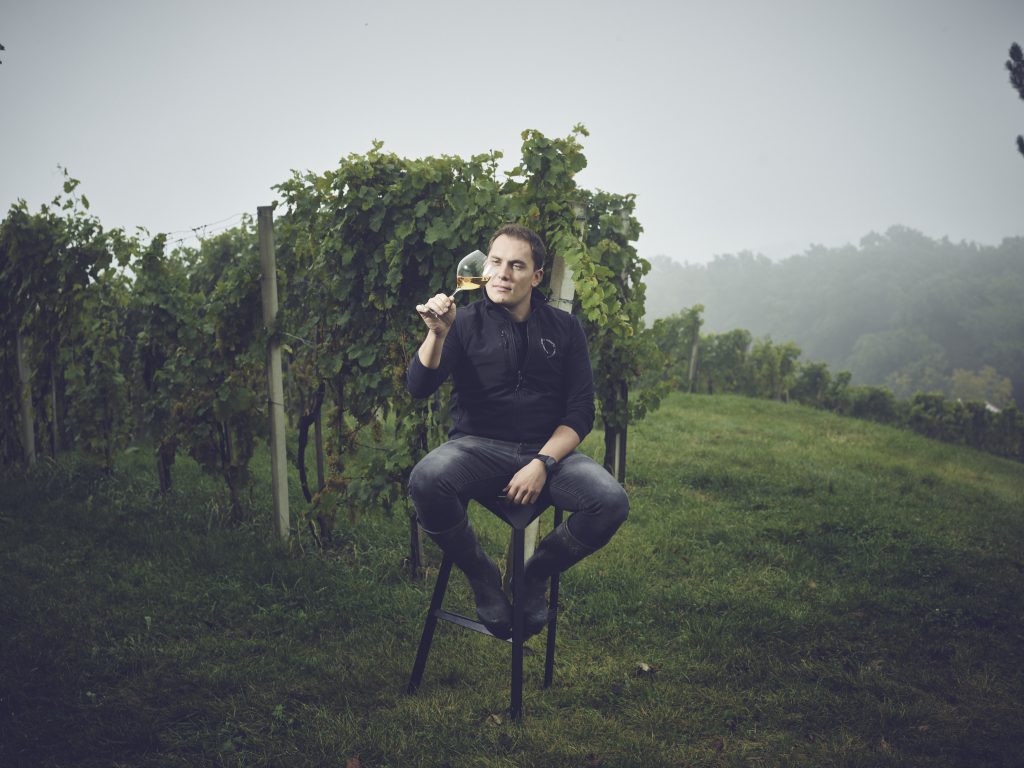 Stefan Krispel auf einem Stuhl mit einem Glas Wein inmitten seiner Weinberge